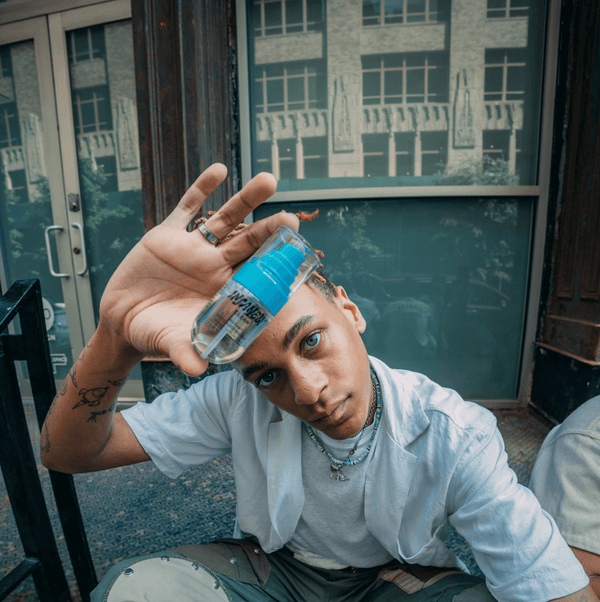 A man sits on the streets of NYC, holding the 42mL Instant Moisturizing Mist from Insanely Clean up to the camera.