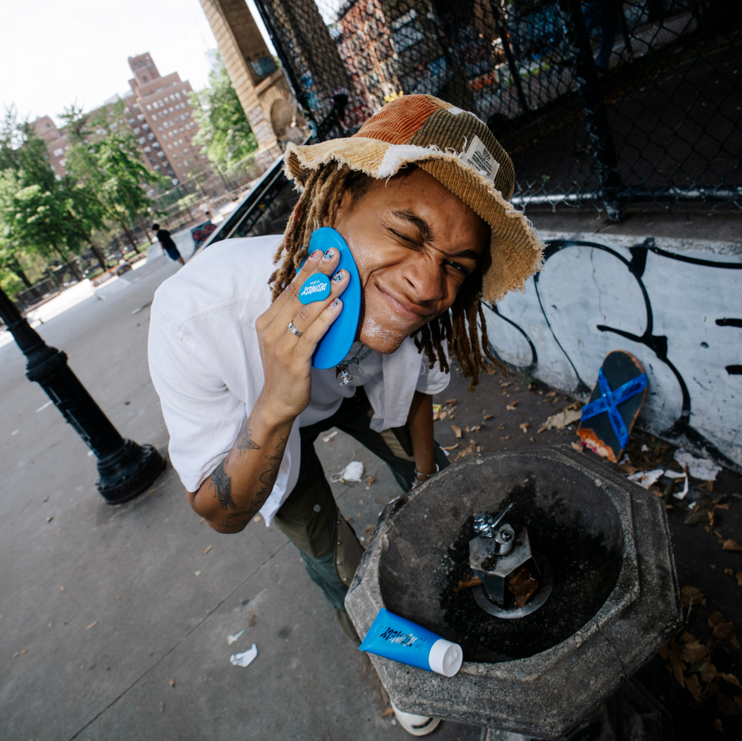 A man scrubbing his face with the Insanely Clean Body Scrubber with the Day 'N' Nite Face Wash resting by a water fountain at a skate park.